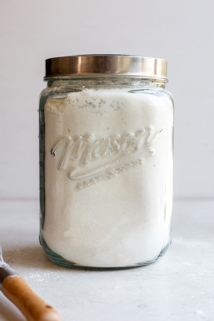 Gluten-Free Bread Flour Blend in a large mason jar with a silver lid. White background, with a whisk in the foreground.