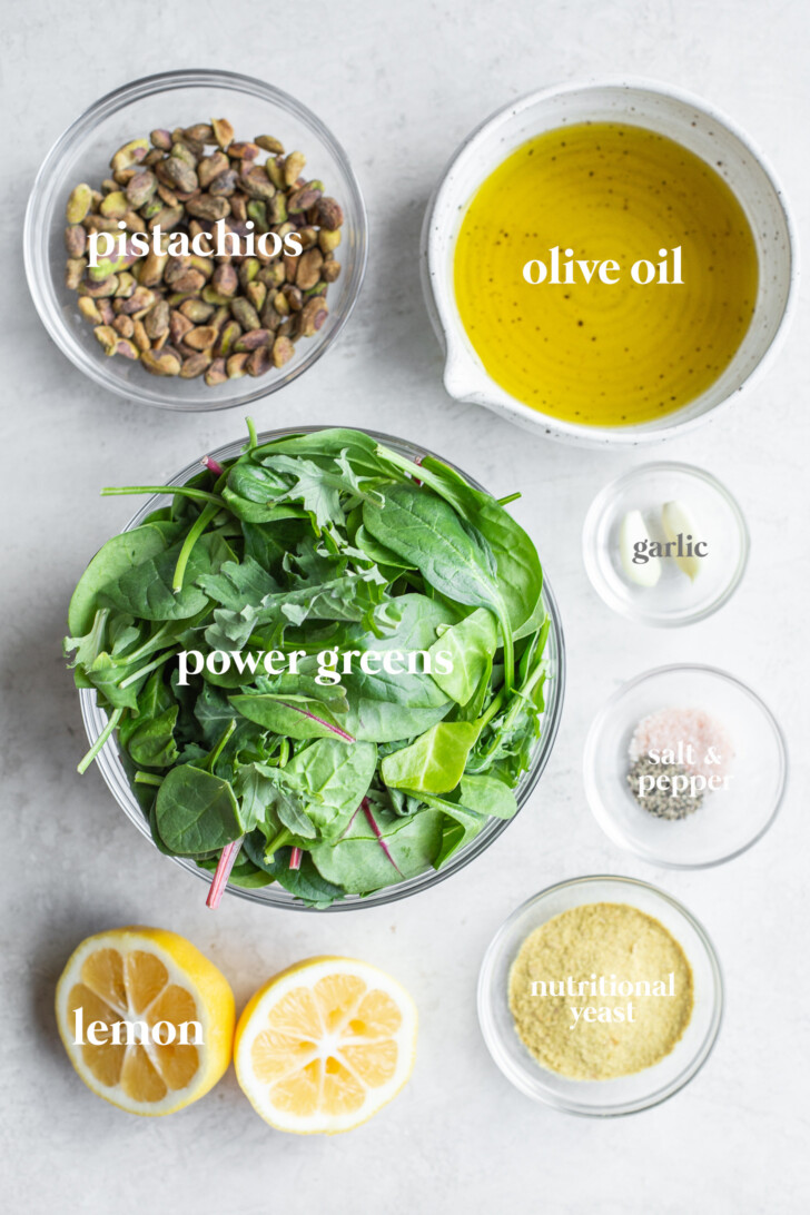 Photo of ingredients for power greens pesto, each in individual glass bowls: power greens, pistachios, olive oil, garlic, salt & pepper, nutritional yeast, and a halved fresh lemon.